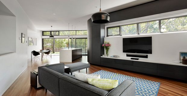 Contemporary open-plan living room with a dark grey sectional sofa, built-in black and white media wall, timber flooring, and a white kitchen island visible behind it. Large horizontal windows bring in natural light and views of trees, while a black pendant light and dark cylindrical architectural feature add contrast.