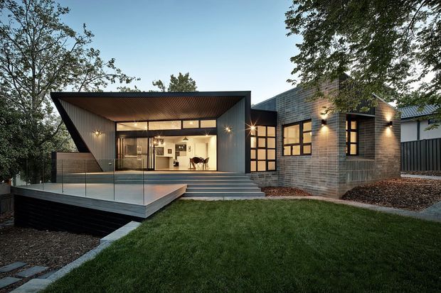 A contemporary single-storey residence photographed at dusk, with angular pavilion-like forms, extensive glazing, a projecting timber-lined roof canopy, and a raised deck overlooking a landscaped lawn. The facade combines dark metal cladding with pale brickwork and warm interior lighting.