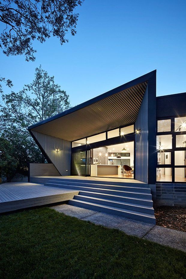 Twilight exterior view of a contemporary single-storey home with a dramatic angular roof canopy, dark metal cladding, extensive black-framed glazing, and a timber-lined soffit. Broad steps lead up to a raised timber deck and open-plan kitchen visible through the illuminated windows. Mature trees and lawn surround the house.