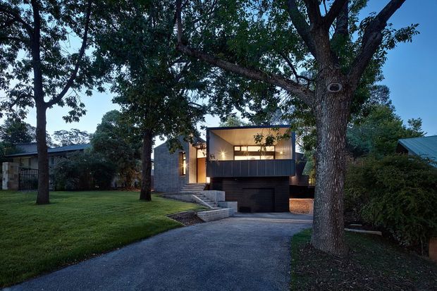 Dusk exterior view of a contemporary two-storey house set among mature trees and landscaped lawns. The home features a flat-roofed upper volume, dark metal or timber-look cladding, expansive warm-lit glazing, a projecting balcony, exposed concrete or masonry side walls, and an integrated garage accessed by a sloping driveway.