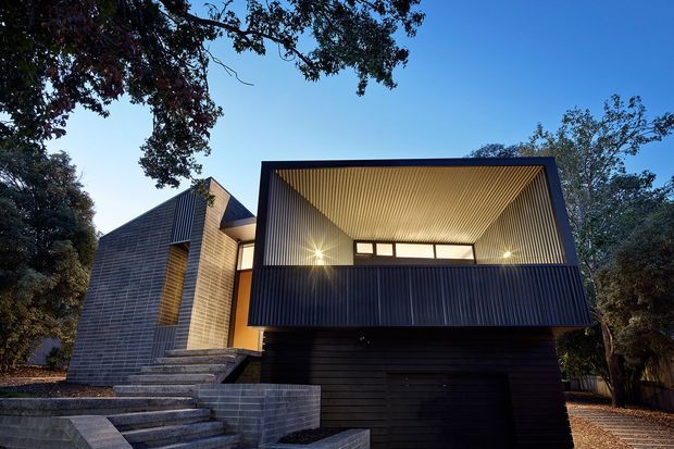 Contemporary multi-level house exterior set among mature trees, featuring dark horizontal cladding, a projecting upper balcony or carport-like volume, a dramatic recessed terrace with a ribbed timber ceiling, exposed masonry walls, and broad stepped entry stairs.
