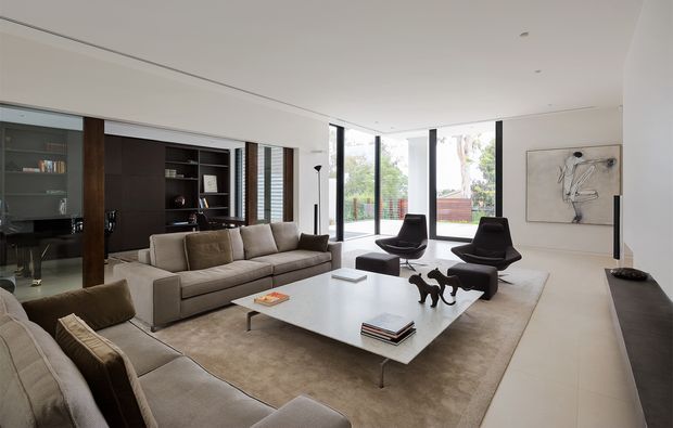 A refined contemporary living room with a neutral palette, large floor-to-ceiling glazed doors, a substantial pale sectional sofa, sculptural dark lounge chairs, and a large white rectangular coffee table. Dark timber built-in cabinetry and shelving line the rear wall, while a minimalist artwork and concealed linear fireplace add understated detail.