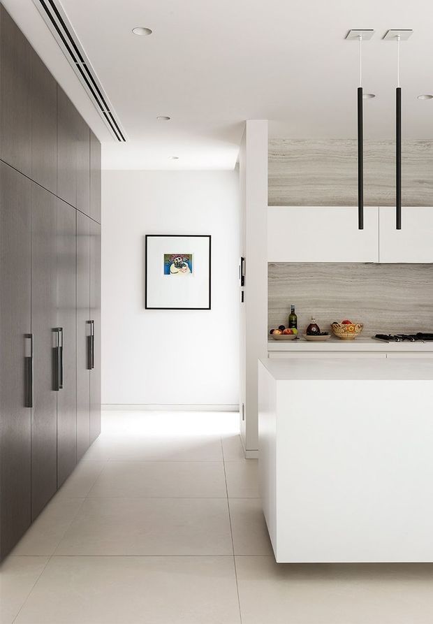 A bright, minimalist contemporary kitchen viewed from an adjacent hallway or circulation space. The kitchen features a large white island, full-height white cabinetry, pale wood-grain cabinetry and splashback surfaces, dark elongated pendant lights, integrated appliances, and a pale tiled floor. A dark built-in storage wall with long vertical handles lines the left side, while a framed artwork hangs on the white hallway wall.