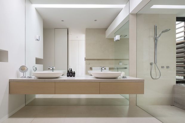 Bright contemporary bathroom with a floating timber vanity, twin white vessel basins, large frameless mirror, pale stone or tiled surfaces, and an open shower with chrome fittings. A high horizontal window with dark louvres provides natural light on the right.