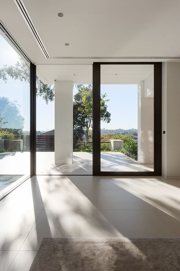 Bright minimalist interior with floor-to-ceiling glazing and a large dark-framed glass door opening onto an outdoor terrace and landscaped view. The space has a white ceiling with recessed downlights, pale tiled flooring, crisp white walls and strong natural sunlight creating shadows across the floor. A partial rug and glimpse of a pool or water feature are visible at the lower left.
