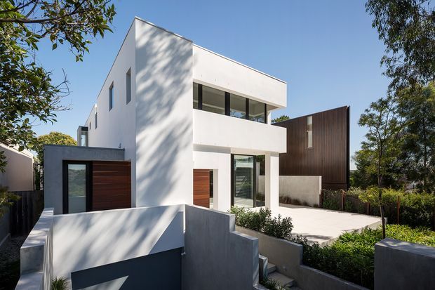 Street-facing view of a contemporary multi-level residence with crisp white rendered walls, projecting rectangular balconies, large dark-framed windows, and timber garage and entry doors. A darker vertically clad neighbouring or secondary volume contrasts with the main façade, while landscaped planting and concrete retaining walls frame the driveway and entry.