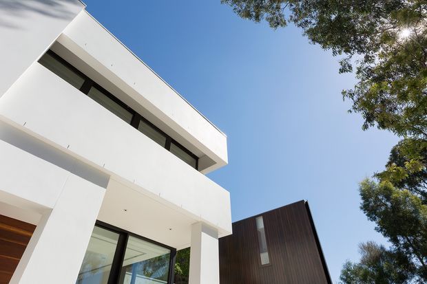 Low-angle exterior view of a contemporary multi-storey residence with crisp white rendered volumes, deep rectangular overhangs, dark-framed glazing, and a contrasting vertically clad dark timber or metal volume. Mature trees frame the building against a clear blue sky.