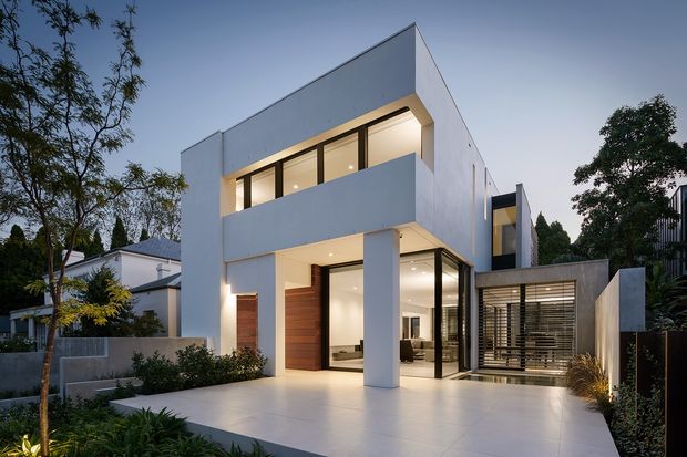 Contemporary two-storey white rendered residence with a crisp geometric flat-roof form, extensive dark-framed glazing, a projecting upper-level balcony, and a broad pale tiled or concrete forecourt. Warm interior lighting, timber entry doors, horizontal privacy screening, landscaped planting, and neighbouring suburban homes are visible.