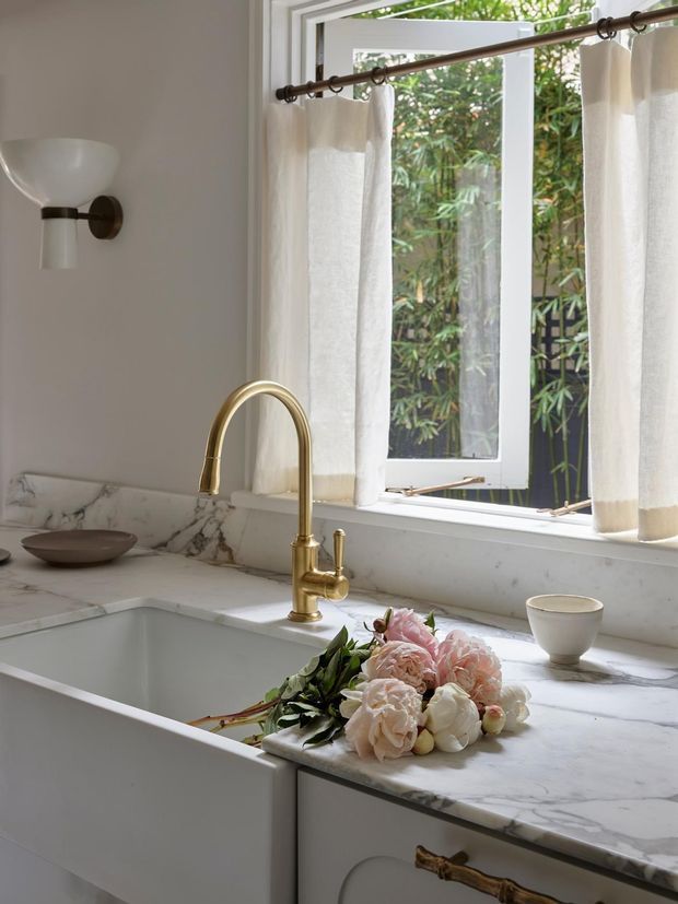A bright, refined kitchen vignette featuring a deep white farmhouse-style sink, white marble benchtops and splashback, pale cabinetry, and a brushed brass gooseneck tap. An open white-framed window with sheer curtains overlooks greenery, while a wall sconce and soft floral arrangement add warmth.