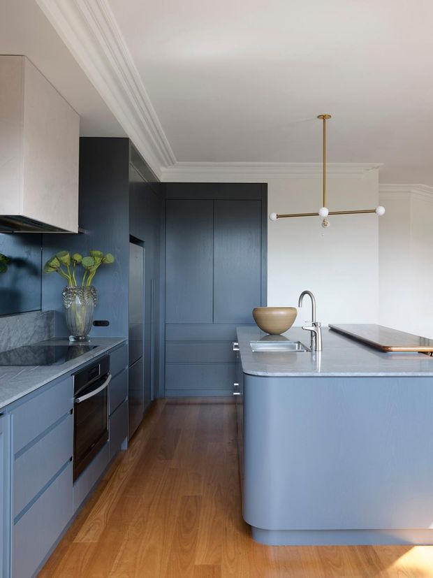 A refined contemporary kitchen featuring deep blue built-in cabinetry, a large curved island with a pale stone benchtop, integrated stainless-steel sink and tapware, timber flooring, white walls with decorative ceiling mouldings, and a sculptural brass-and-opal pendant light. The space has a sophisticated blue-and-white palette with warm timber accents.