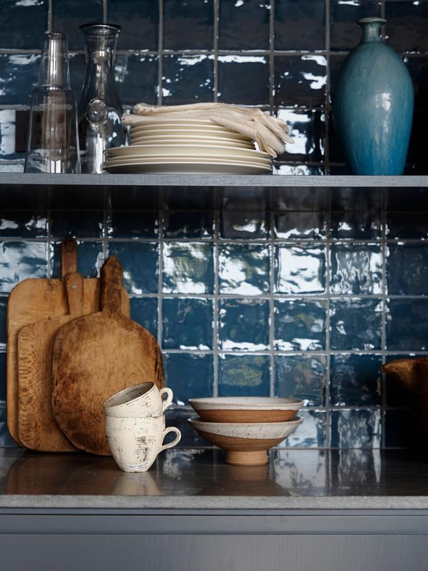 Close-up view of a styled kitchen shelving and benchtop vignette. Glossy, irregular dark blue ceramic tiles form the splashback, with open grey shelves displaying stacked cream plates, glass vessels, a blue ceramic bottle, wooden chopping boards, rustic cups and bowls. The lower cabinetry has a muted grey finish, creating a moody, artisanal atmosphere.