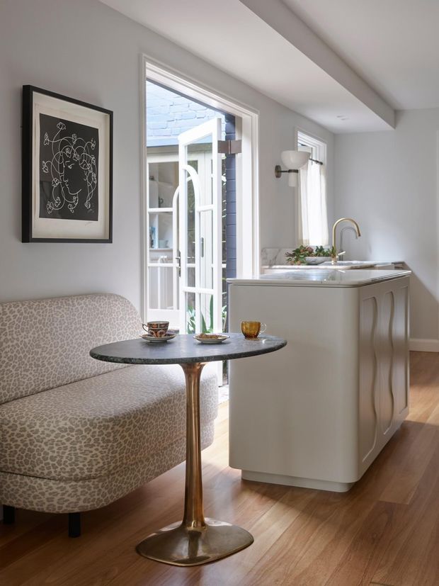 A bright, refined open-plan interior featuring a compact kitchen island with a pale stone benchtop and sculptural white cabinetry, paired with a curved upholstered banquette and small round pedestal table. White-framed glazing and an open glazed door bring in daylight, while warm timber flooring and restrained wall lighting create an inviting atmosphere.