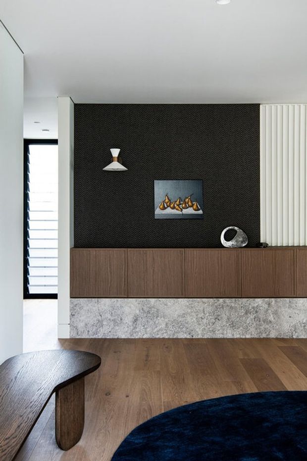 A refined contemporary living room or entry area featuring a full-width dark chevron-patterned feature wall, floating timber cabinetry, a textured grey stone base, and vertical white wall panelling. Warm timber flooring, sculptural decor, artwork, a curved timber table, and a dark blue rug create a sophisticated, high-contrast interior.