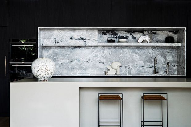 A sophisticated contemporary kitchen featuring a large white island with integrated seating, dark timber cabinetry, and a dramatic grey-and-white marble backsplash. Open shelving displays ceramics, while black metal fixtures and bar stools add contrast.