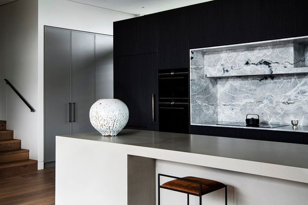 Contemporary open-plan kitchen with a large pale island and integrated seating, dark timber cabinetry, grey stone benchtops, and a dramatic veined stone splashback. A sculptural pendant light hangs above the island, with a timber staircase and tall grey pantry cabinetry visible behind.