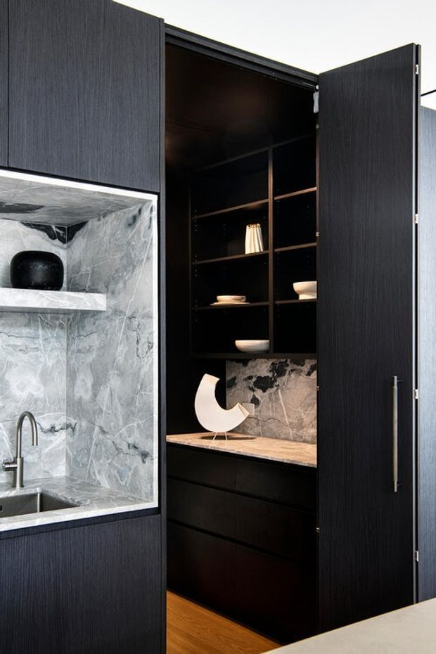 Close-up interior view of a sophisticated dark kitchen or butler's pantry joinery installation. Tall black timber-grain cabinetry conceals an open pantry cupboard with adjustable shelving, decorative crockery, drawers and a warm timber benchtop. Adjacent is a compact sink area lined in dramatic grey veined stone, with a slim stone shelf and brushed metal tap. The palette is predominantly black, charcoal, grey marble and warm wood, creating a refined, high-contrast contemporary look.