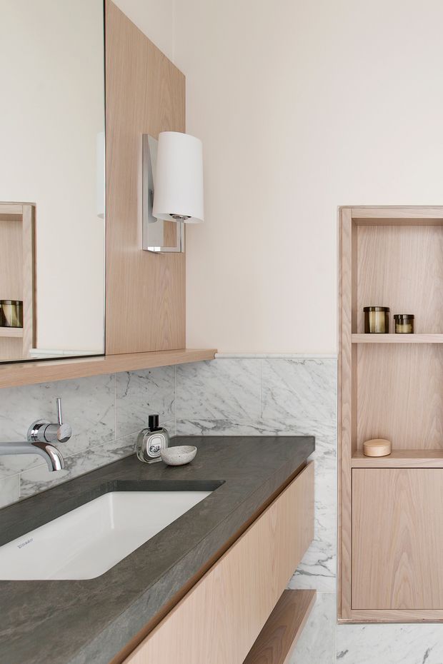 Close-up view of a refined bathroom vanity featuring a white integrated basin, dark grey stone benchtop, pale natural timber cabinetry and open shelving, and white marble-look wall and floor tiles. A large mirror and cylindrical wall sconce sit above the vanity, with minimal toiletries and neutral styling.
