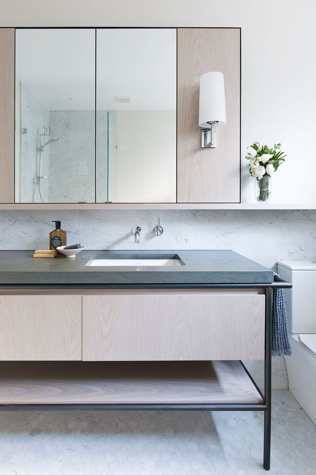 Bright, refined contemporary bathroom featuring a wall-mounted pale timber vanity with an open lower shelf, dark stone benchtop and integrated basin, marble-look wall surfaces, small light floor tiles, mirrored upper cabinetry, glass shower enclosure, and a sculptural wall sconce. A toilet and folded towel are visible at right.