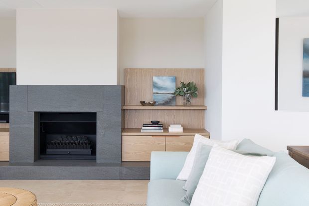 A bright contemporary living room featuring a substantial grey stone fireplace with a dark inset firebox, pale timber built-in cabinetry and display shelving, and a light upholstered sofa in the foreground. White walls, restrained artwork, and neutral furnishings create a clean, calm interior.