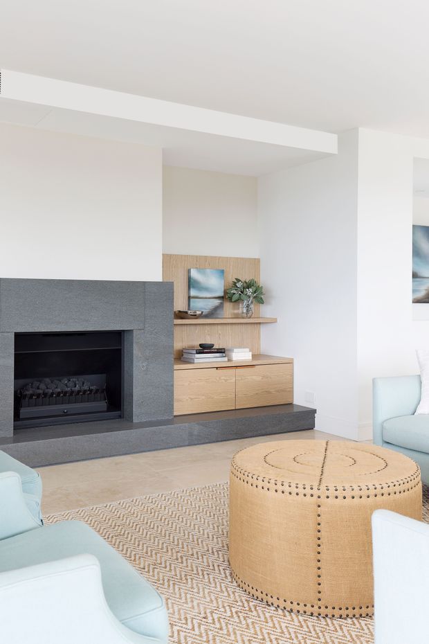 Bright contemporary living room with a built-in dark grey stone fireplace and raised hearth, light timber entertainment cabinetry and shelving, white walls, pale upholstered armchairs, and a large round tan upholstered ottoman over a patterned neutral rug.