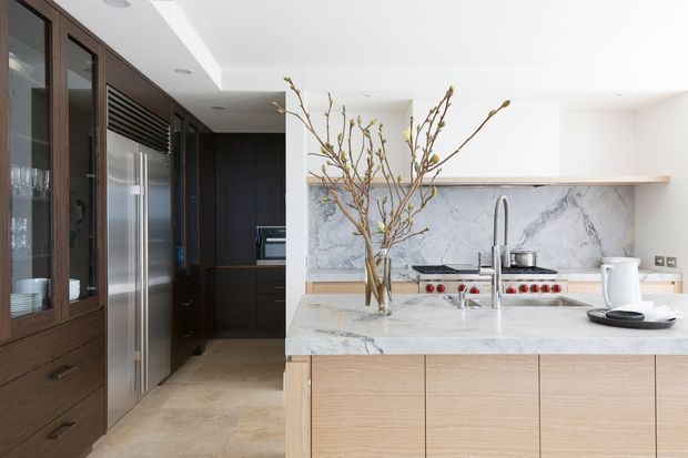 Bright contemporary kitchen with a large marble island, integrated stainless-steel appliances, pale timber cabinetry, dark timber pantry joinery, and a sculptural branch arrangement. The space has a clean, refined material palette with strong natural light and concealed ceiling lighting.