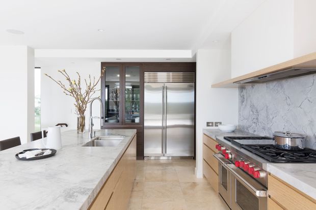 Bright contemporary kitchen with a large marble island, light timber cabinetry, stainless-steel refrigerator and range cooker, and a marble backsplash. Dark-framed glazed cabinetry and abundant white surfaces create a clean, refined look.