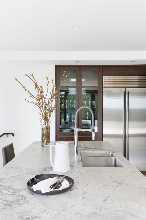 Bright contemporary kitchen interior featuring a large veined marble island with an integrated stainless-steel sink and chrome gooseneck faucet. Dark timber-framed glass cabinetry and a large stainless-steel refrigerator form the rear wall, with white walls and ceiling creating a clean, refined look. A dining chair, decorative branches, and tableware are visible around the island.