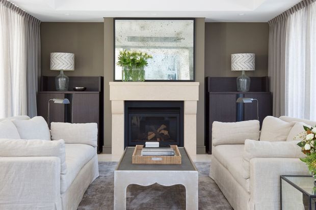 A refined living room arranged symmetrically around a central fireplace. Two light upholstered sofas face each other across a sculptural coffee table, with dark built-in or freestanding cabinetry, matching lamps, full-height curtains, and a large framed artwork or mirror above the mantel. The palette is neutral and warm, with layered textures and balanced contemporary-classic styling.