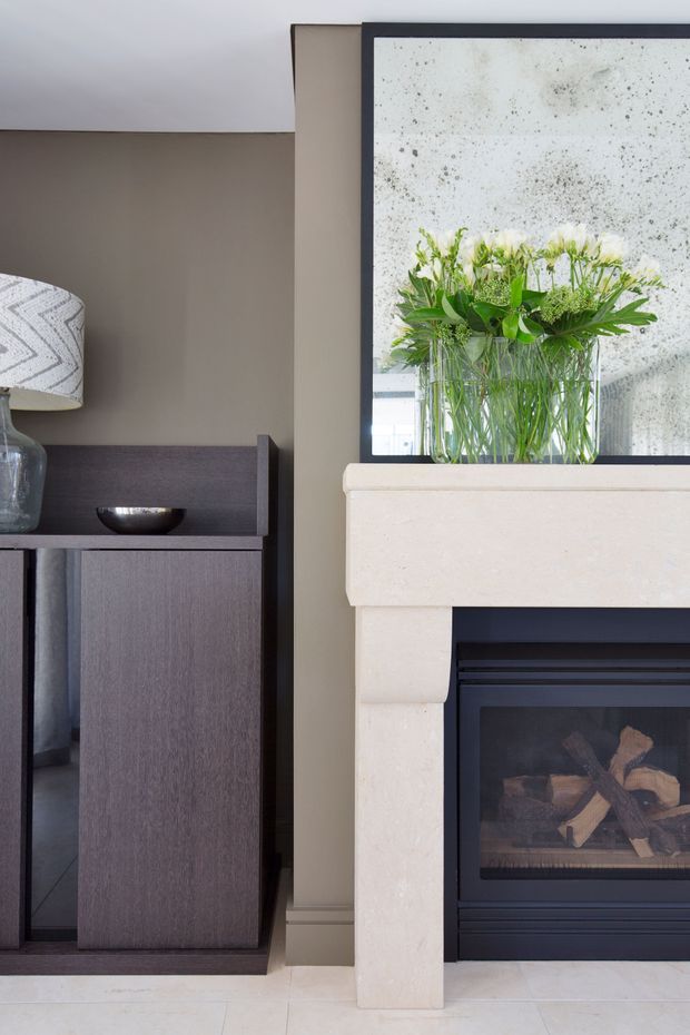 Close-up interior view of a refined living room featuring a pale stone fireplace surround with a black inset gas fire, a dark timber built-in cabinet, neutral taupe wall panelling, and a large dark-framed mirror above the mantel with a glass vase of greenery.