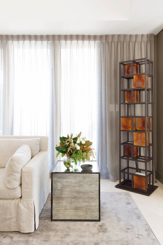 Bright, refined living room corner with a cream upholstered sofa, sheer floor-to-ceiling curtains, a textured neutral rug, and a mirrored or metallic side table. A tall dark metal open shelving unit displays warm amber decorative glass objects, adding contrast and a luxurious focal point. A floral arrangement and small bowl sit on the side table.