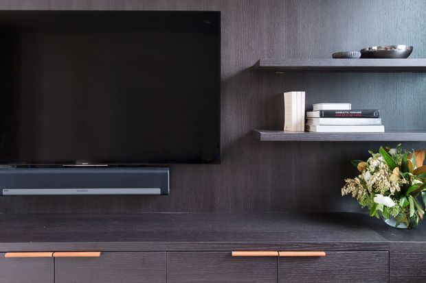 Close-up of a dark, built-in media wall featuring a large flat-screen television, soundbar, floating display shelves with books and decorative objects, and a low dark timber entertainment cabinet with copper-toned handles. A floral arrangement adds a natural accent.