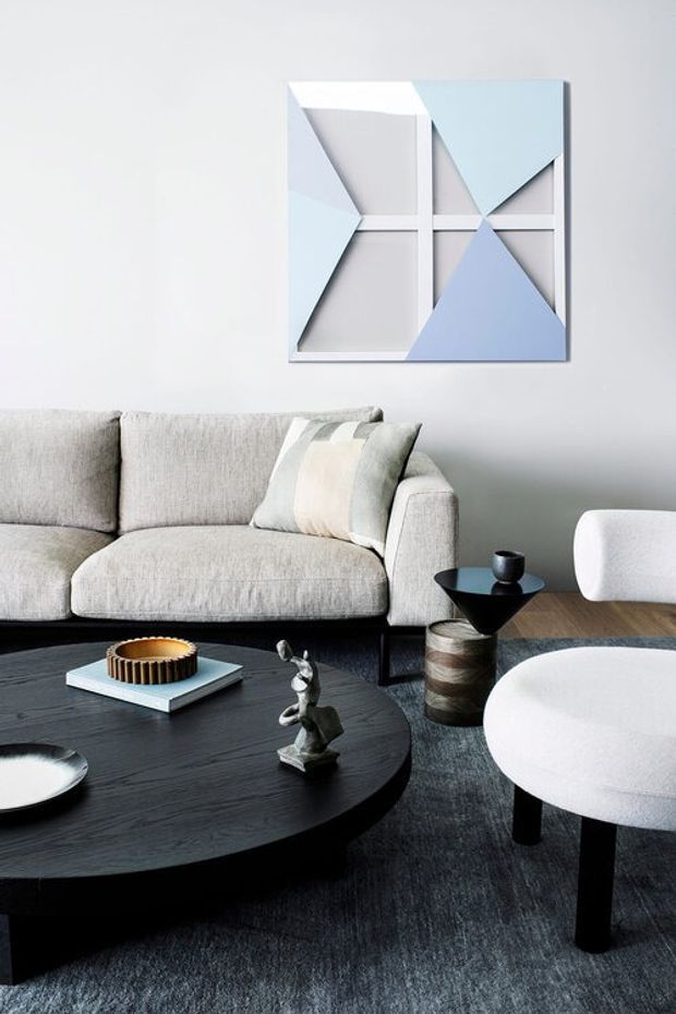 A refined contemporary living room featuring a pale upholstered sofa, a white rounded accent chair, a large black oval coffee table, a small sculptural side table, and a muted blue-grey rug. Geometric blue, white, and grey wall art provides a subtle feature above the sofa, while neutral upholstery and dark timber furniture create a calm, sophisticated setting.