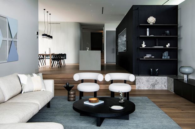 A refined open-plan contemporary living space with a pale sectional sofa, sculptural white lounge chairs, a low black oval coffee table, and a muted blue-grey rug over warm timber flooring. A dining area and compact kitchen are visible behind the living zone, alongside dramatic black built-in shelving and cabinetry with a stone-clad base. Vertical wall panelling and modern pendant lighting add architectural interest.