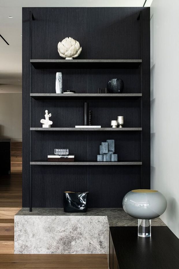 A refined interior vignette featuring a tall, dark timber built-in shelving unit with open shelves displaying sculptural decorative objects. The shelving sits above a dark stone or textured benchtop, with a black console surface and a large pale stone coffee table in the foreground. Warm timber flooring and a neutral white wall balance the moody joinery, while a sculptural table lamp adds a soft contemporary accent.