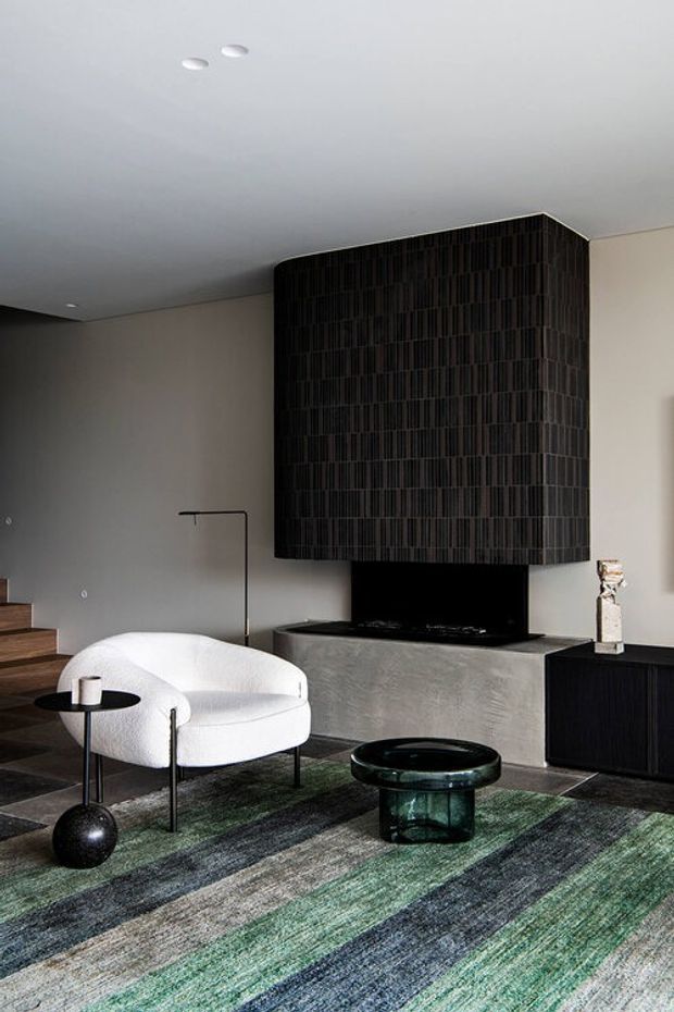 A sophisticated contemporary living room with a sculptural white lounge chair, dark geometric coffee table, black sideboard, and a large fireplace framed by a dramatic dark tiled feature wall. The room includes a grey concrete-look hearth, neutral plaster walls, timber stair treads, recessed ceiling lights, and a large striped green, grey, and charcoal rug. A slender black floor lamp and decorative objects complete the refined, moody interior.