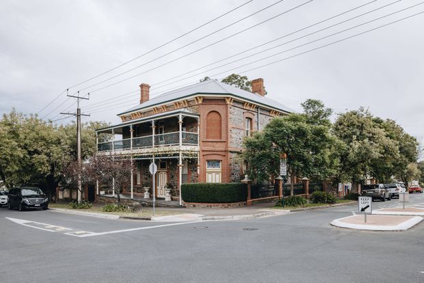 Street-level exterior view of a two-storey late-19th-century brick corner building with a wraparound verandah, decorative rendered trim, tall chimneys, arched upper window detailing, and timber-framed windows and doors. The building sits on a landscaped urban intersection with overhead utility wires and mature trees.