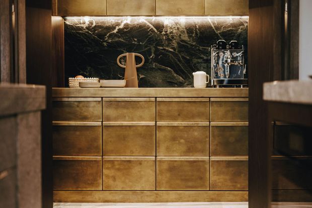 A compact, moody drinks and coffee station framed by dark timber openings. The built-in cabinetry and overhead cupboards have a warm brushed brass or bronze finish, with a dark veined stone splashback, integrated warm LED lighting, and a countertop holding an espresso machine, carafe, cups, and pastries.