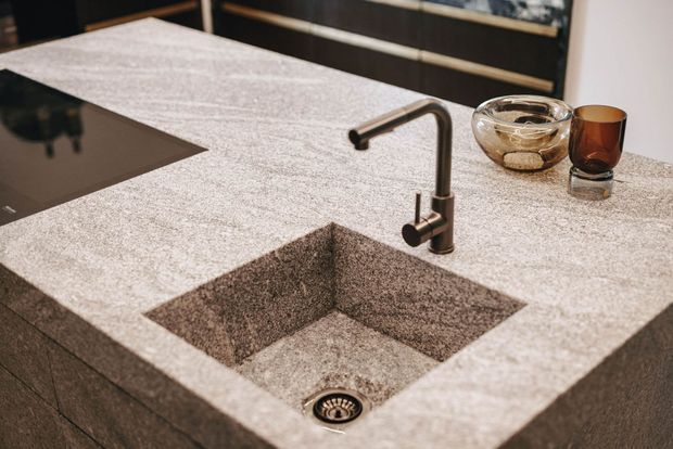 Close-up view of a contemporary kitchen benchtop featuring an integrated square stone sink, dark pull-out mixer tap, black induction cooktop, and dark cabinetry in the background. The benchtop has a speckled grey-beige stone finish, complemented by smoked glassware and a glass bowl.