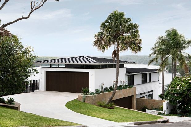 Contemporary multi-level residence on a sloping site, featuring crisp white rendered walls, dark standing-seam metal roofing, a large dark timber-toned garage door, and horizontal cladding. The house is surrounded by manicured lawns and tropical palms, with a water view visible beyond the property.