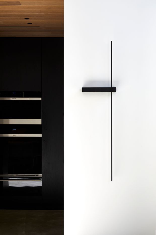 Close-up of a minimalist built-in entertainment or appliance wall. A tall black joinery unit with integrated dark appliances occupies the left side beneath a warm timber ceiling or overhead panel, while a clean white wall on the right features a slim black linear wall detail or handle. The composition is stark, refined, and highly minimal.