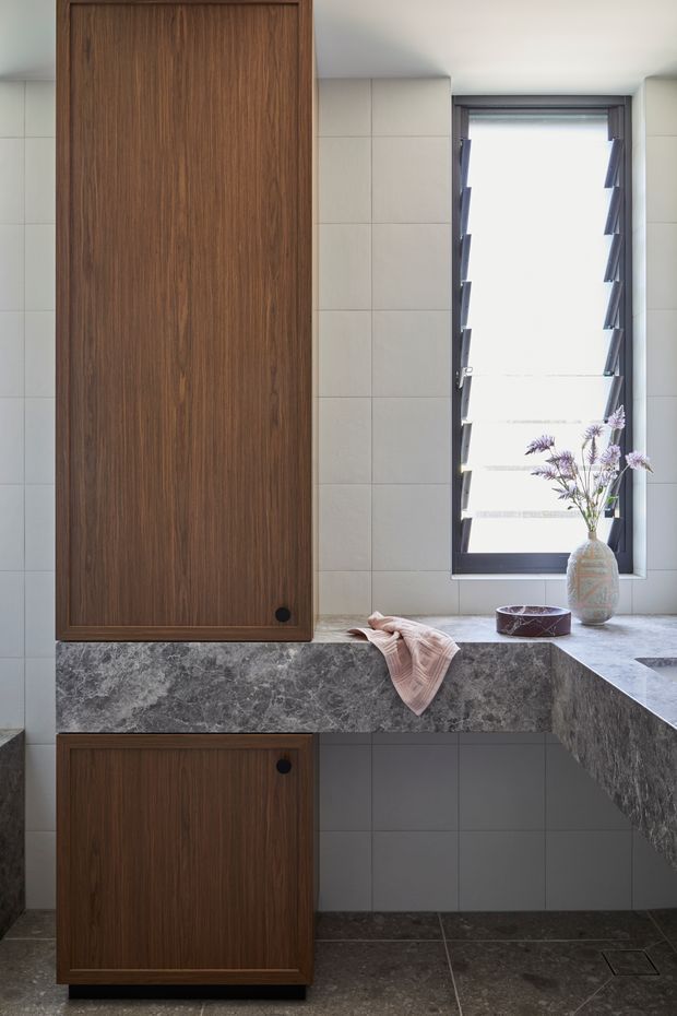 Contemporary bathroom vanity area featuring dark timber cabinetry, large grey stone benchtops, pale square wall tiles, dark floor tiles, and a black-framed louvred window. A vase with flowers and a folded towel add restrained decorative touches.