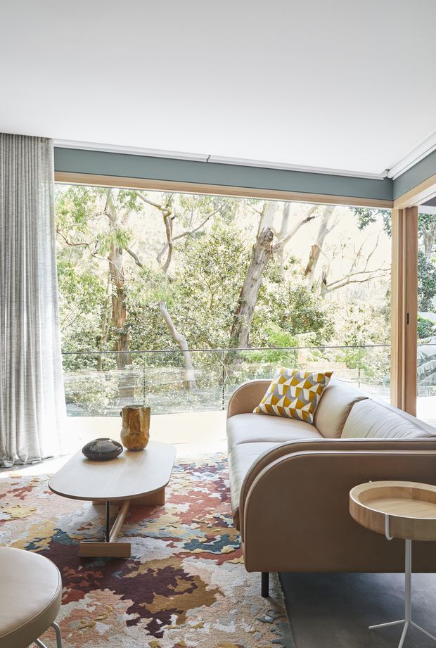 Bright, refined living room with a curved tan sofa and matching rounded coffee table arranged on a richly patterned area rug. Full-height glazing and timber-framed corner windows open to leafy trees, while sheer curtains soften the natural light. A sculptural side table, decorative vessels, and a geometric mustard-and-white cushion add mid-century character.