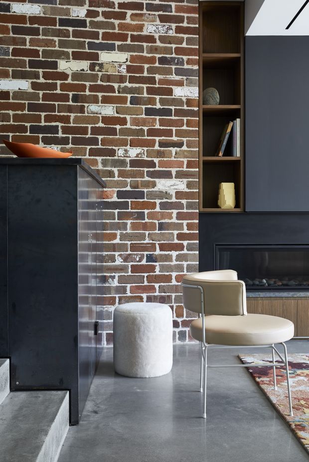 A refined interior vignette featuring an exposed red-and-white brick wall, dark built-in cabinetry with open shelving and a fireplace, a polished grey concrete floor, and a sculptural cream lounge chair beside a small white upholstered stool. A colourful patterned rug and minimalist decor add contrast to the industrial material palette.