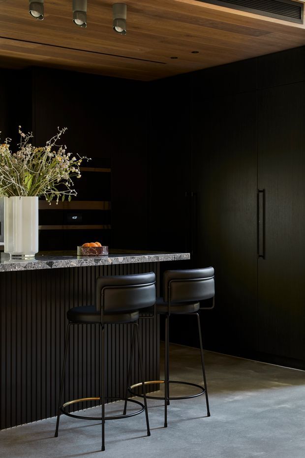 A dark, sophisticated kitchen or home bar area with black vertically fluted cabinetry, a substantial stone counter, and two black bar stools. The ceiling is clad in warm timber with recessed and track lighting, while a large black built-in cabinet occupies the rear wall. Decorative branches in a tall vase and a small bowl sit on the counter.