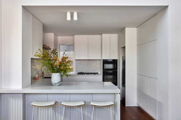 Bright, contemporary white kitchen with a large island peninsula, three stools, integrated cabinetry, a gas cooktop, and built-in black appliances. The room has a clean minimalist fit-out, pale stone-like surfaces, white vertical-panel island detailing, and dark timber flooring. A vase of branches provides a small natural accent.