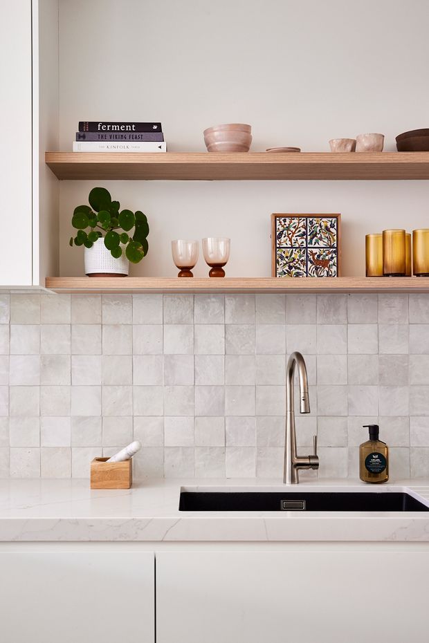 A close-up view of a refined kitchen sink area featuring pale square tiled splashback, white cabinetry, a light stone benchtop, brushed metal tapware, and open timber shelving displaying books, ceramics, glassware, artwork, and a potted plant.