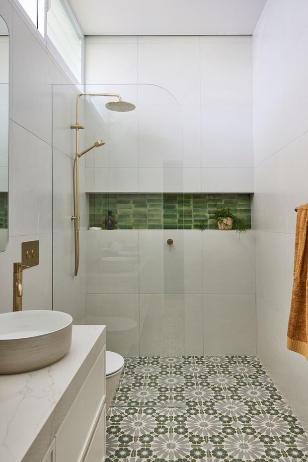 Bright contemporary bathroom with large white wall tiles, a clear glass walk-in shower and brushed brass shower fittings. A green glazed-tile recessed niche adds colour above the wall-hung toilet, while patterned encaustic-style floor tiles create a decorative feature. The room includes a white vessel basin on a pale vanity, a narrow high window, and a warm ochre towel.