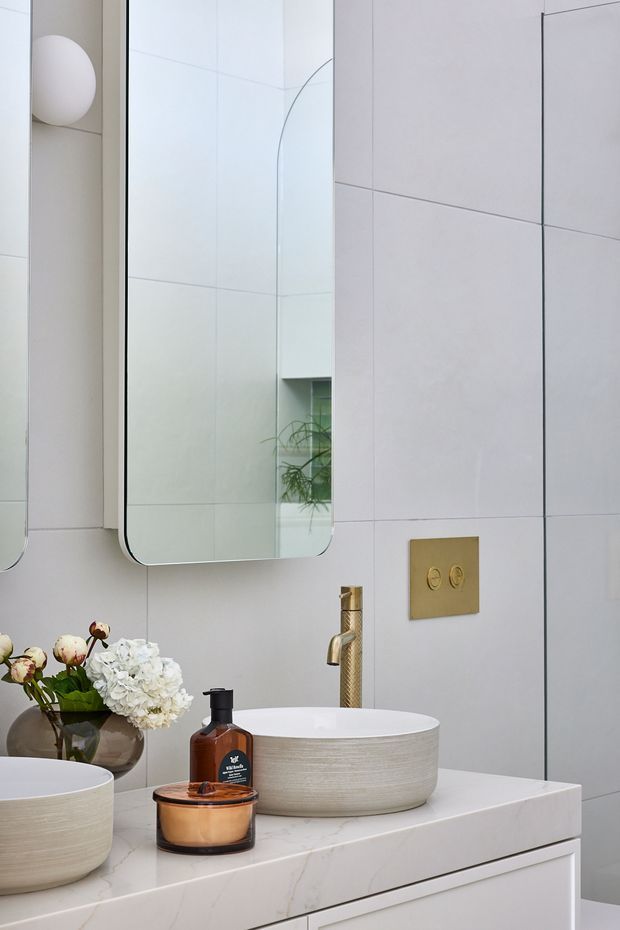 A refined contemporary bathroom vanity with two round vessel basins on a pale stone benchtop, brushed gold tapware and matching flush plate, large mirrored cabinetry, and oversized pale wall tiles. Soft floral styling and warm metallic details add contrast to the light, minimal space.