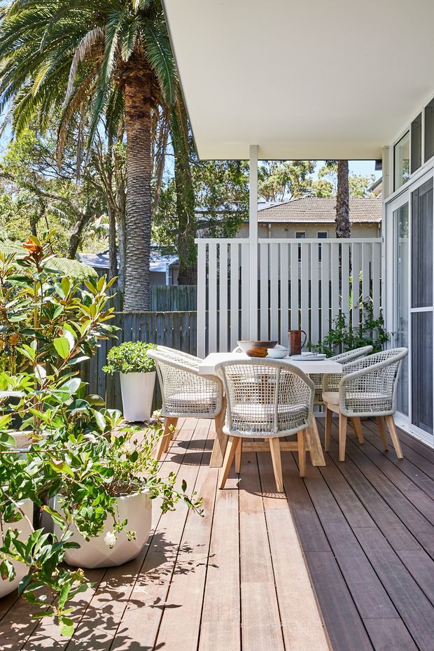 Sunlit outdoor dining terrace with timber decking, a white vertical-slat privacy screen and covered roof, furnished with a light outdoor dining table and woven chairs. Large potted plants and mature palms create a leafy suburban setting, with a glazed house facade along one side.