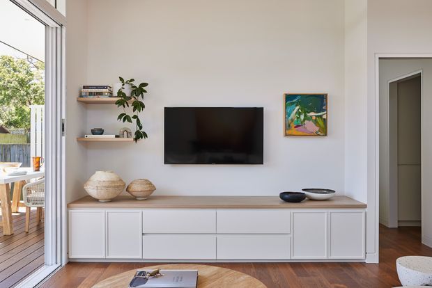 Bright contemporary living room centred on a wall-mounted television and low built-in white media cabinetry with a light timber benchtop. Open shelving displays books and decor, while an open glazed door connects to a timber outdoor dining area. White walls, timber flooring, artwork and restrained furnishings create a clean, warm interior.